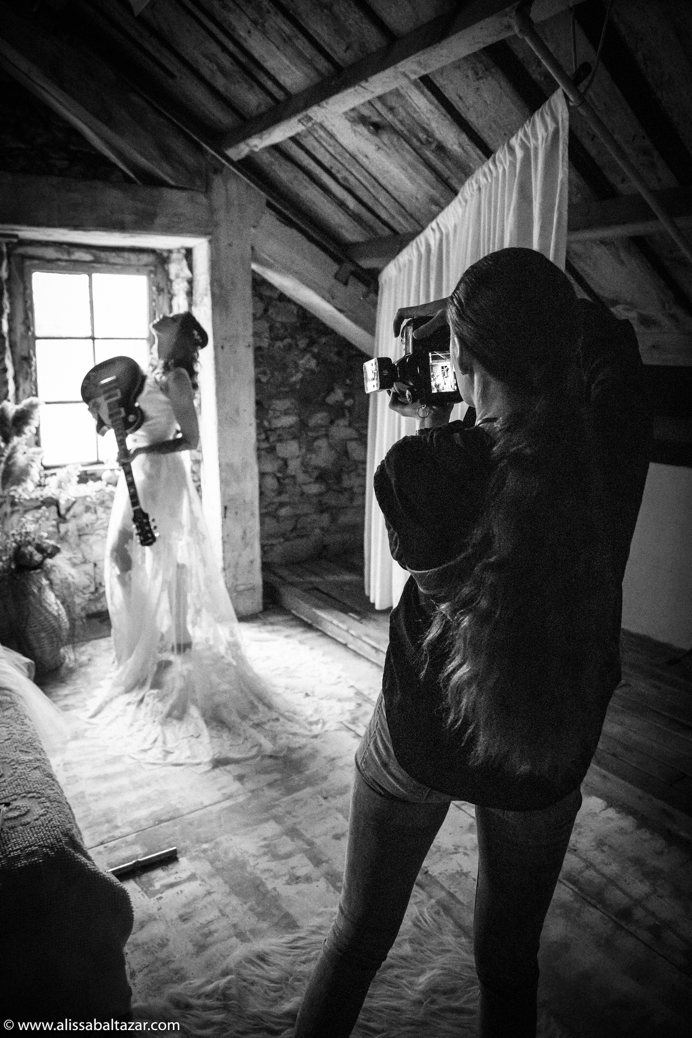 Looking from behind at Alissa Baltazar photographer as she takes a rock and roll wedding picture featuring a bride with a guitar in an antique backlit window