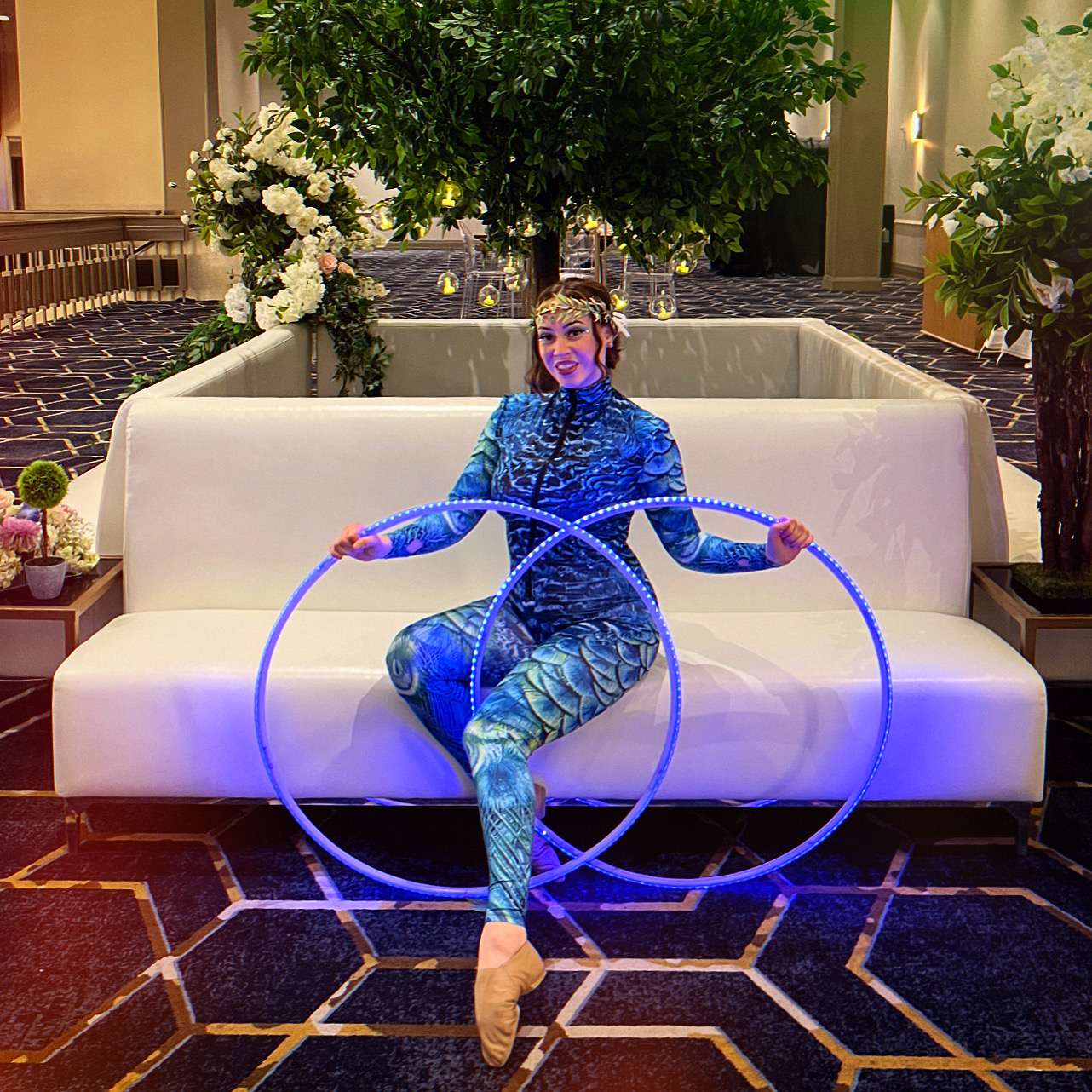 Lyza moon poses on a white couch with LED hula hoops, they are lit up with blue lights,  wearing a blue costume in a banquet wall